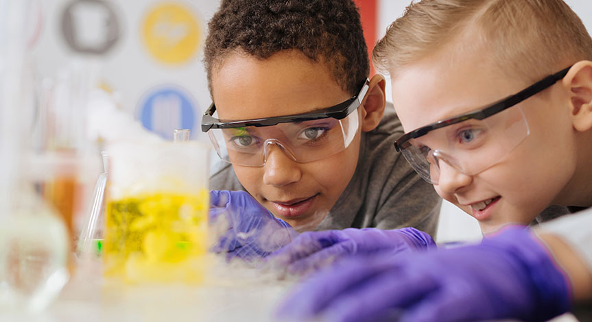 boys in safety goggles leaning closely to the table and watching chemical reaction going on in the beaker