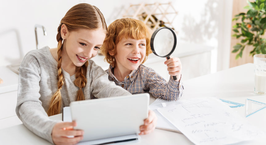 Boy looking through a magnifying glass while girl next to him looks at her computer while doing a Mad Science virtual class